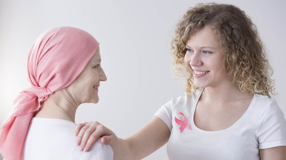 Mulher sorridente apoiando outra mulher em tratamento, ambas com o laço rosa